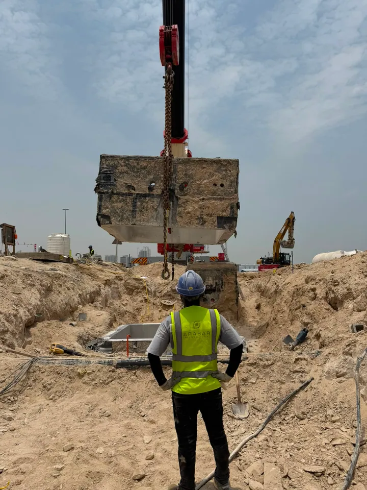A crane lifting a large concrete chamber section above an excavated work area