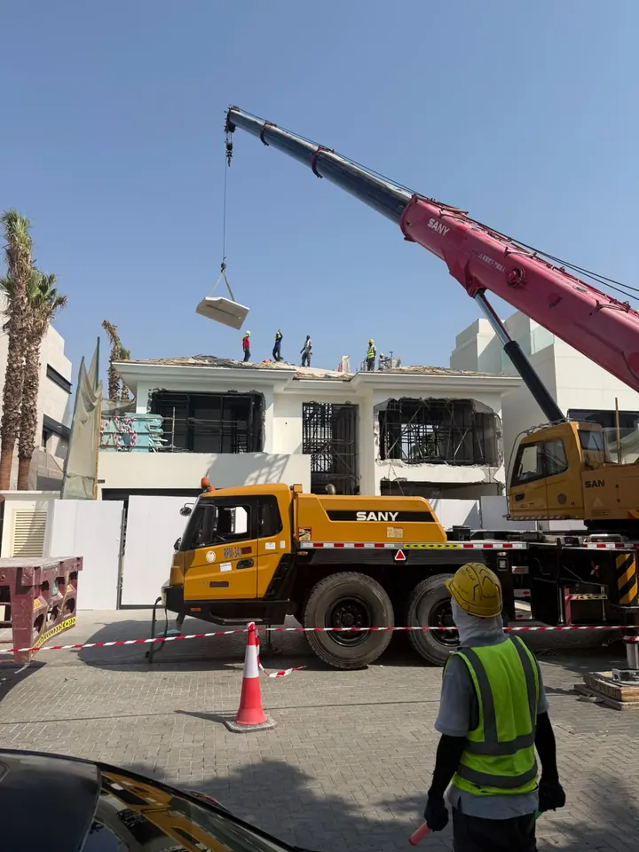 A mobile crane lifting a rectangular concrete section above a villa during Arasar works